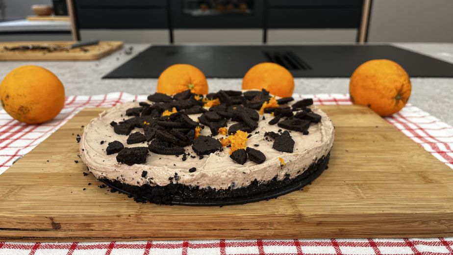 Ein fertig zubereiteter „Black Cheesecake“ auf einem Holzbrett, garniert mit groben dunklen Keksstücken und frischem Orangenabrieb. Der Kuchen zeigt den deutlichen Kontrast zwischen dem dunklen Oreo-Keksboden und der hellen Creme-Füllung, während im Hintergrund ganze Orangen die herbe Note des Desserts unterstreichen.