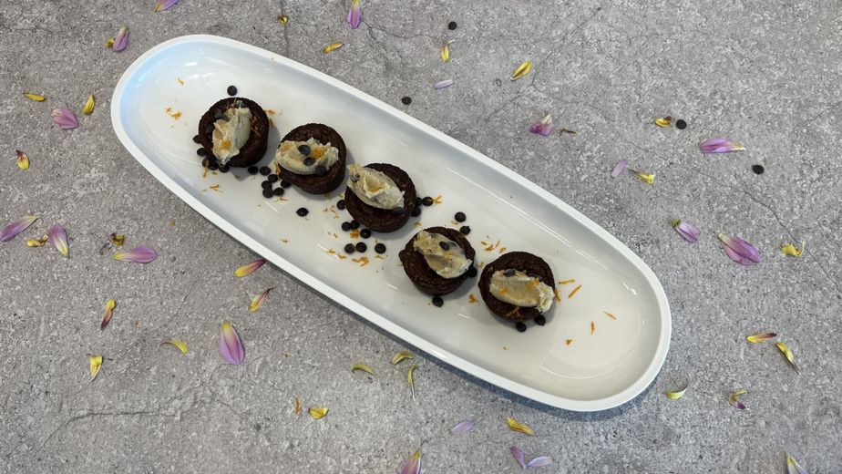 Fünf fertige Brownies sind auf einer länglichen, weißen Servierplatte angerichtet. Auf jedem Brownie sitzt eine Nocke aus Cookie Dough, die mit Schokotropfen bestreut ist. Weitere Schokotropfen und dekorative Blütenblätter sind um die Brownies auf der Platte und dem grauen Untergrund verteilt.