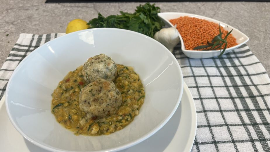 Servierfertiger, cremiger Linseneintopf in einer weißen Schüssel, getoppt mit zwei hausgemachten Semmelknödeln. Im Hintergrund sind die frischen Zutaten wie rote Linsen, Petersilie, Knoblauch und eine Zitrone dekorativ auf einem karierten Küchentuch angerichtet.