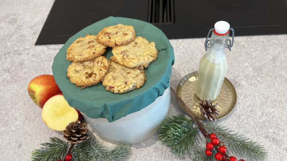 Goldbraun gebackene Bratapfel-Cookies, angerichtet auf einer mit grünem Tuch bedeckten Dose, neben einer Bügelflasche mit cremigem Vanillekipferl-Likör. Das Arrangement auf der Arbeitsplatte wird durch frische Äpfel, Tannenzweige und Zapfen weihnachtlich dekoriert.