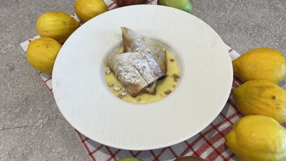 Zwei Stücke goldbrauner Mini-Apfelstrudel, mit Puderzucker bestäubt, serviert in einem tiefen weißen Teller mit cremiger Vanillesauce und gehackten Walnüssen. Der Teller ist auf einem Küchentuch angerichtet und von frischen Äpfeln und Zitronen umgeben, die die Hauptzutaten des Rezepts widerspiegeln.
