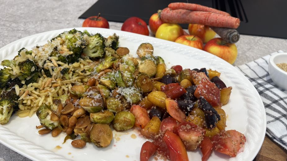 Ein weißer Teller mit drei Portionen Ofengemüse: gerösteter Brokkoli mit Mandelsplittern und Parmesan, Kohlsprossen mit Erdnüssen sowie bunte Karottenstücke mit Sesam. Im Hintergrund sind frische Äpfel und ganze Karotten auf einer Arbeitsplatte zu sehen.