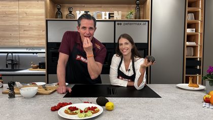 Tanja Pfaffeneder und Philipp von „Philipp bewegt“ präsentieren in einer modernen Küche das Trend-Rezept Cloud Bread. Im Vordergrund ist ein Teller mit dem fertig belegten, proteinreichen Brot mit Avocado und Tomaten zu sehen, während die beiden Gastgeber mit frischen Zutaten posieren.