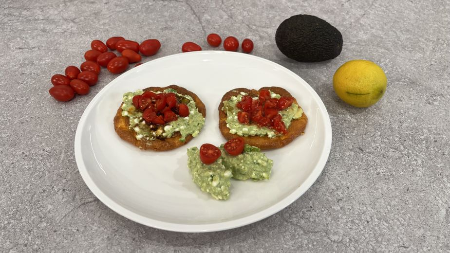 Zwei goldbraun gebackene Cloud-Bread-Fladen auf einem weißen Teller, großzügig belegt mit einer Avocado-Hüttenkäse-Mischung und gewürfelten Kirschtomaten. Im Hintergrund sind eine ganze Avocado, eine Zitrone und lose Kirschtomaten als Zutaten auf einer grauen Arbeitsfläche angerichtet.