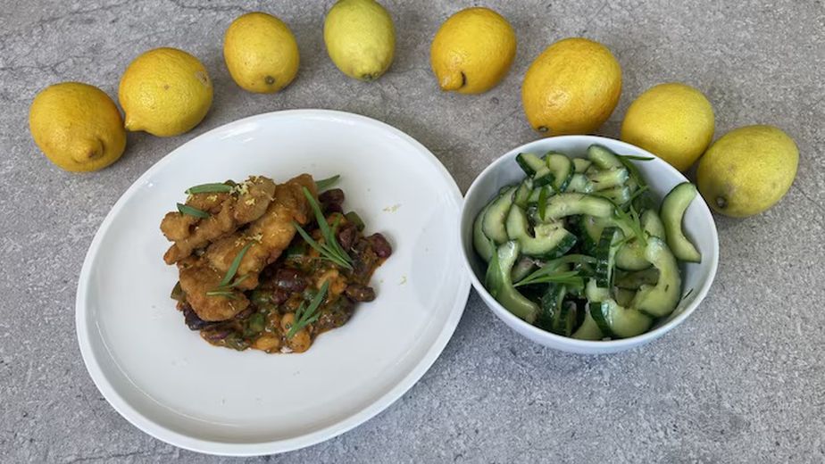 Fertig angerichtetes Backhendl auf Bohnengemüse, serviert auf einem weißen Teller neben einer Schüssel Gurkensalat. Im Hintergrund sind frische Zitronen als Dekoration platziert.
