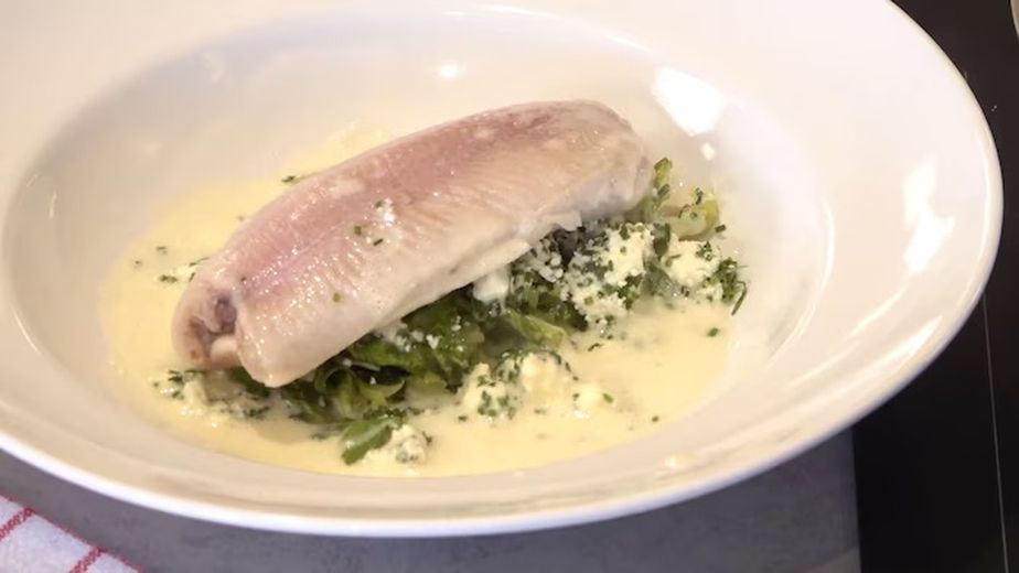 Ein pochiertes Forellenfilet, angerichtet in einer weißen Schüssel auf einem Bett aus Lauch-Romanasalat. Das Gericht ist mit selbstgemachtem Frischkäse bestreut und mit einer hellen, cremigen Molke-Sauce übergossen.