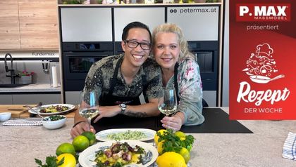 Peter Lehner und Schauspielerin Susanna Hirschler stehen lächelnd in einer modernen Küche nebeneinander und halten jeweils ein Glas Weißwein. Vor ihnen auf dem Küchentresen sind fertig angerichtete mediterrane Gerichte mit Seeteufel und Oktopus sowie Zitronen und Limetten zu sehen.