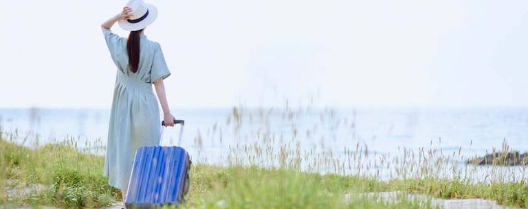Femme de dos regarde la mer, une valise à la main