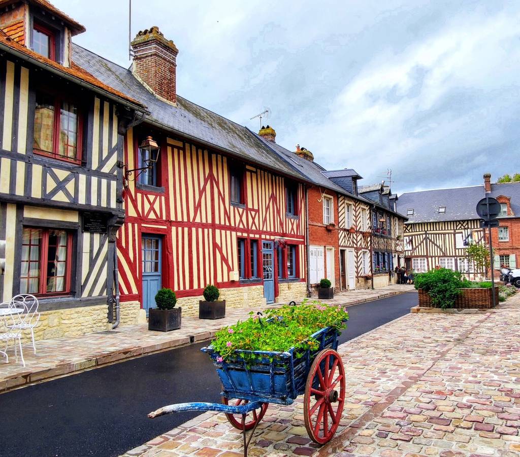 Une rue pittoresque pavée, bordée de maisons à colombages colorées, où trône une charrette bleue ornée de plantes vertes.
