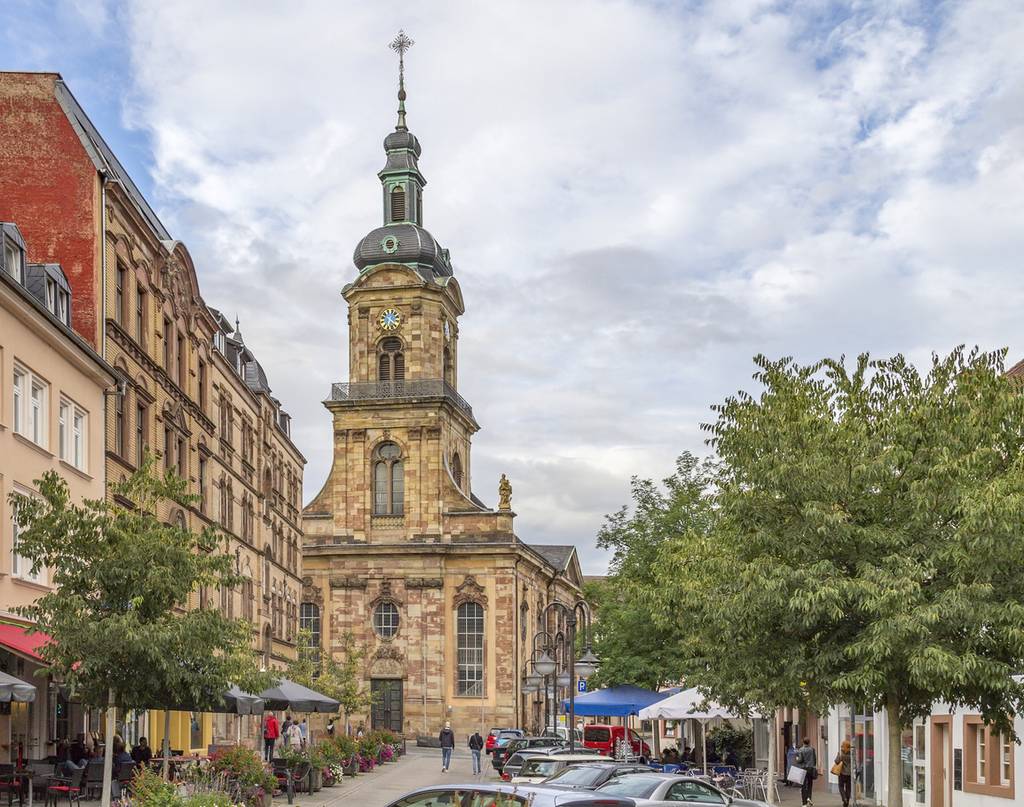 Historische Kirche mit hohem Uhrturm an einer von Bäumen und Gebäuden gesäumten Straße, mit Fußgängern und parkenden Autos unter einem teilweise bewölkten Himmel
