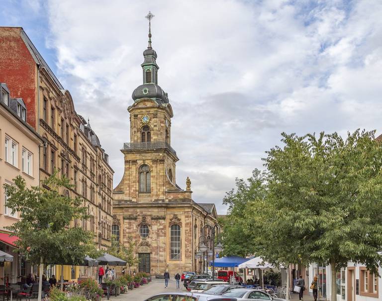 Historische Kirche mit hohem Uhrturm an einer von Bäumen und Gebäuden gesäumten Straße, mit Fußgängern und parkenden Autos unter einem teilweise bewölkten Himmel