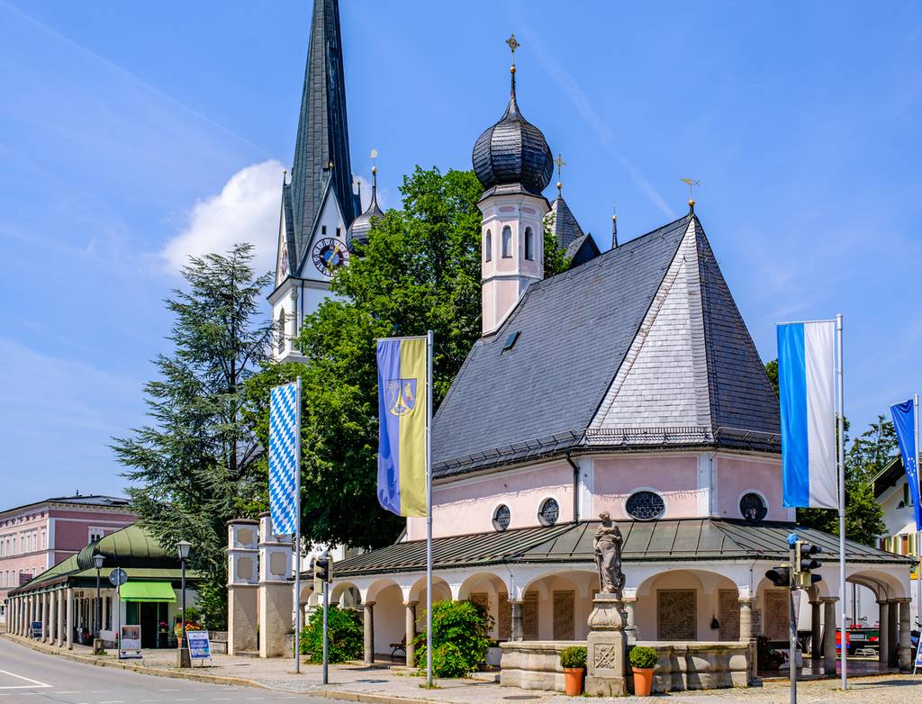 Eine idyllische europäische Kirche mit einem Kirchturm und einem Kuppelturm, umgeben von Flaggen und Bäumen, vor einem klaren blauen Himmel.