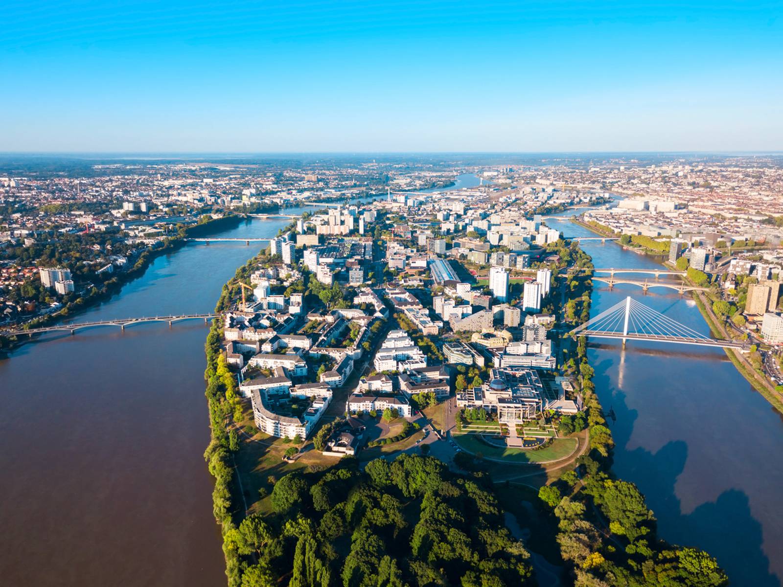 Aerial view of nantes with a river dividing urban areas, bridges connecting the land, and greenery along the riverbanks under a clear blue sky.