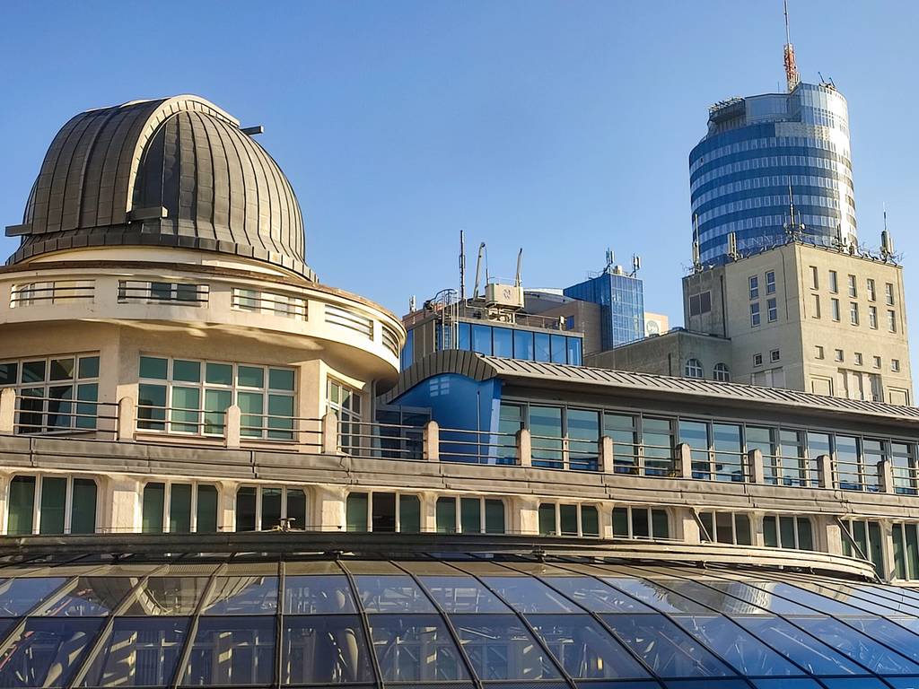 Stadtansicht mit einer Kuppel-Sternwarte, einem modernen Glasgebäude und einem hohen zylindrischen Turm vor klarem, blauem Himmel
