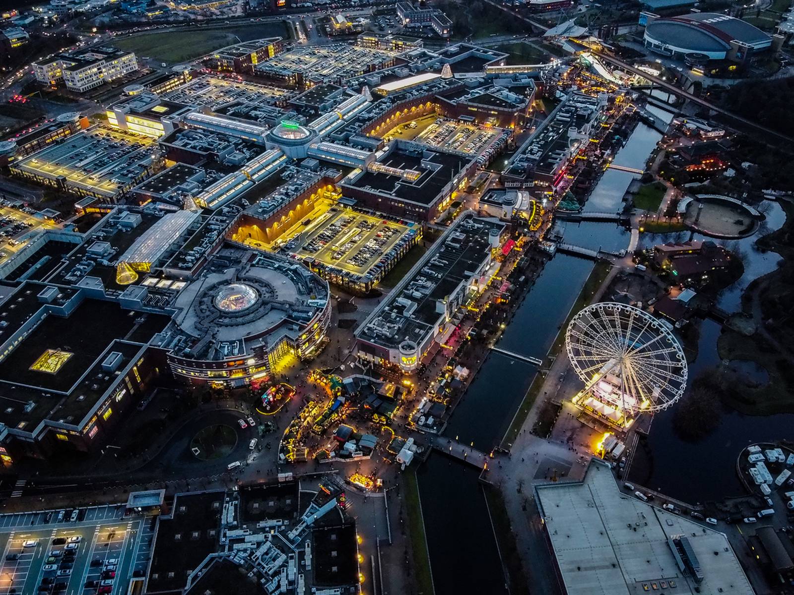 Luftaufnahme einer hell erleuchteten Stadtlandschaft bei Nacht mit einem großen Einkaufszentrum, einem Riesenrad und einem Kanal, der durch das Gebiet verläuft
