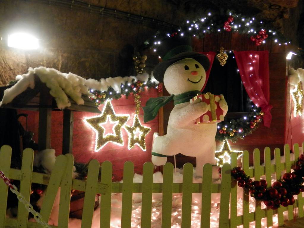 Festlicher Schneemann mit einem Geschenk in der Hand, umgeben von Lichtern, Sternen und weihnachtlichen Dekorationen, hinter einem Lattenzaun mit Schnee und Girlanden.