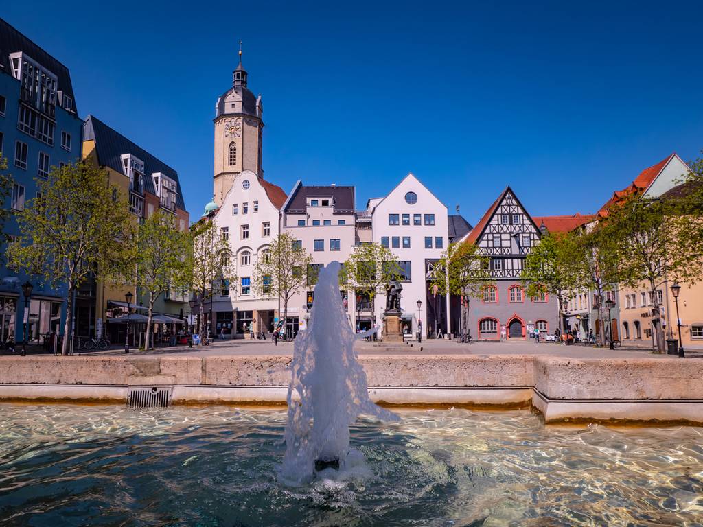 „Historischer Marktplatz mit zentralem Brunnen, umgeben von farbenfrohen Gebäuden und einem Uhrturm unter klarem, blauem Himmel