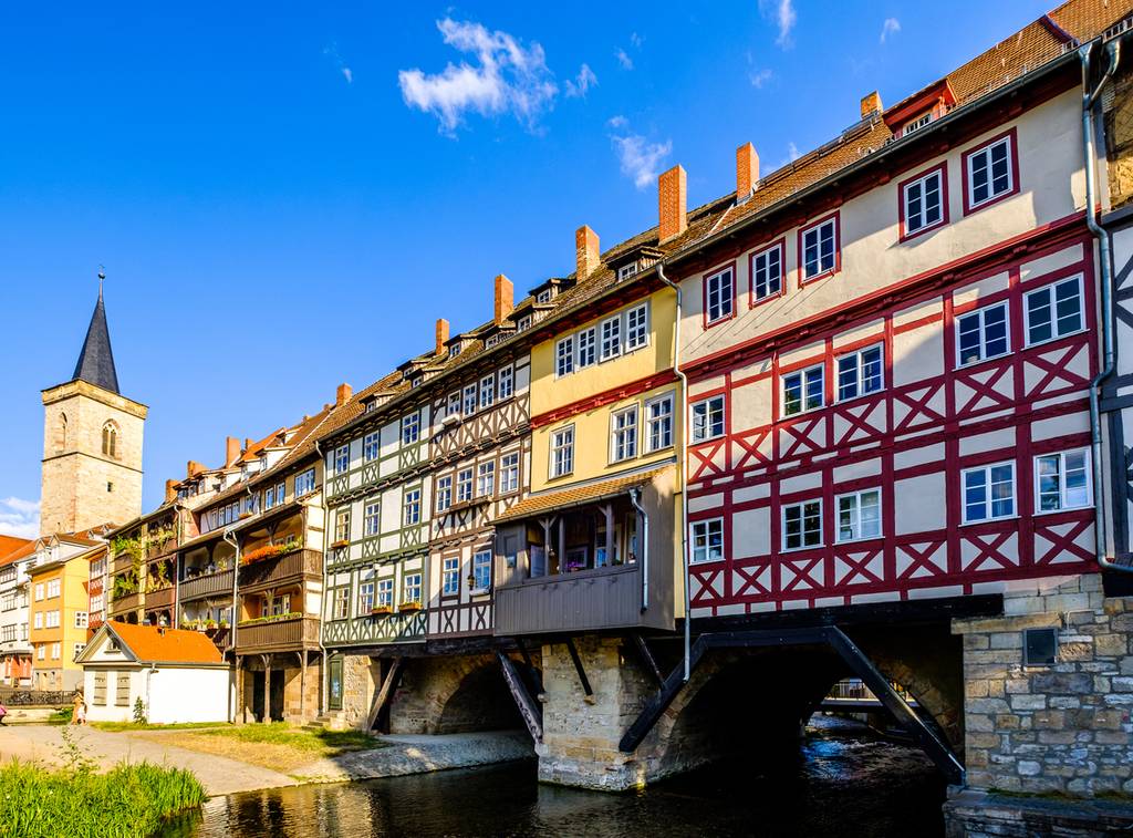Eine Reihe bunter Fachwerkhäuser auf einer Brücke über einem Fluss, mit einem Kirchturm im Hintergrund unter klarem, blauem Himmel