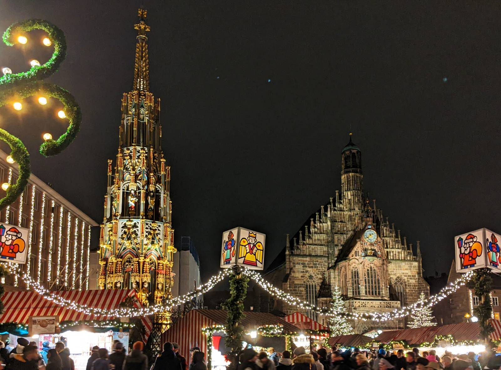 Festlicher Weihnachtsmarkt bei Nacht mit beleuchteten Ständen, einem hohen, verzierten Turm und einem großen historischen Gebäude im Hintergrund