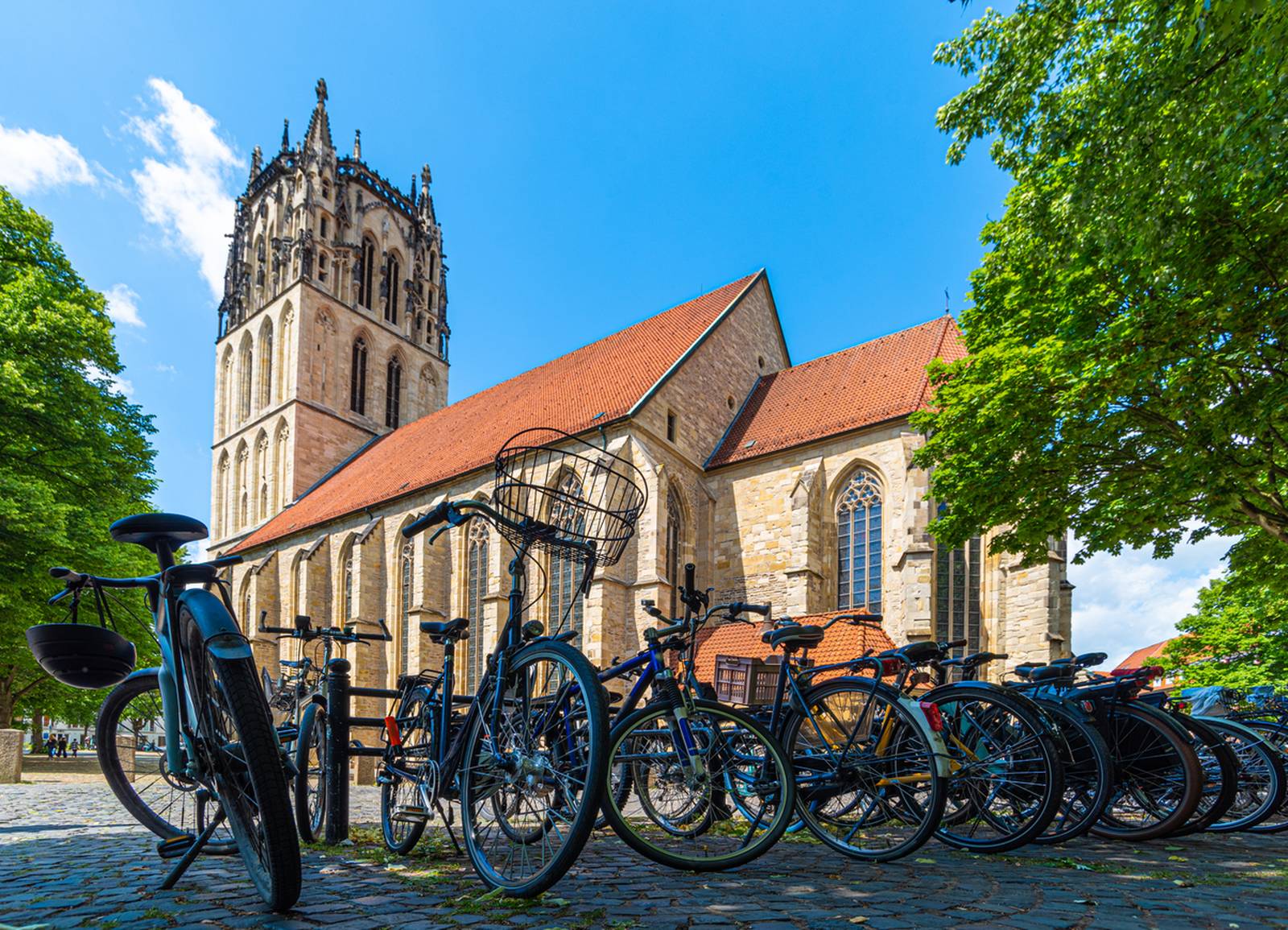 Eine historische Kirche mit hohem Turm steht an einem sonnigen Tag hinter einer Reihe geparkter Fahrräder, umgeben von üppig grünen Bäumen
