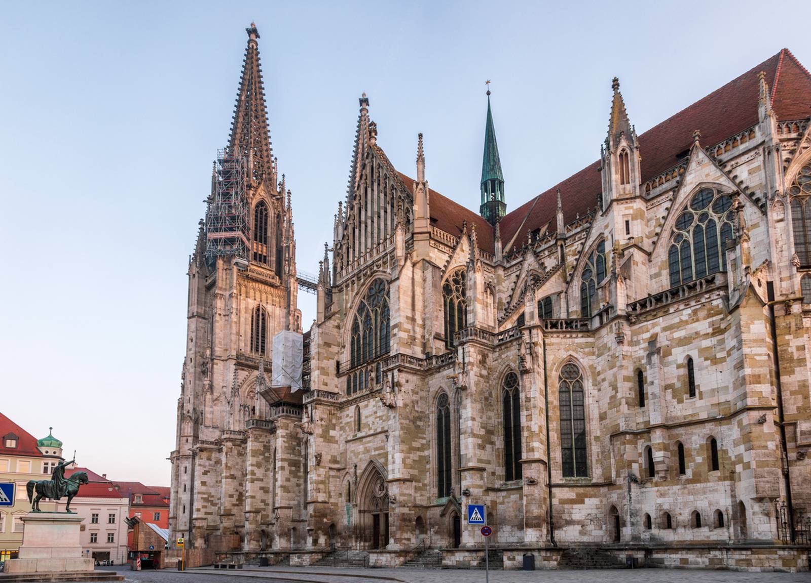 Gotische Kathedrale mit hohen Türmen und detaillierter Steinmetzarbeit vor klarem Himmel, mit einer Statue und Gebäuden im Hintergrund