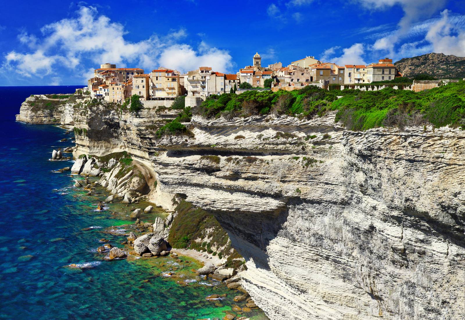 Clifftop Bonifacio with buildings overlooking a turquoise sea, set against a blue sky with scattered clouds.