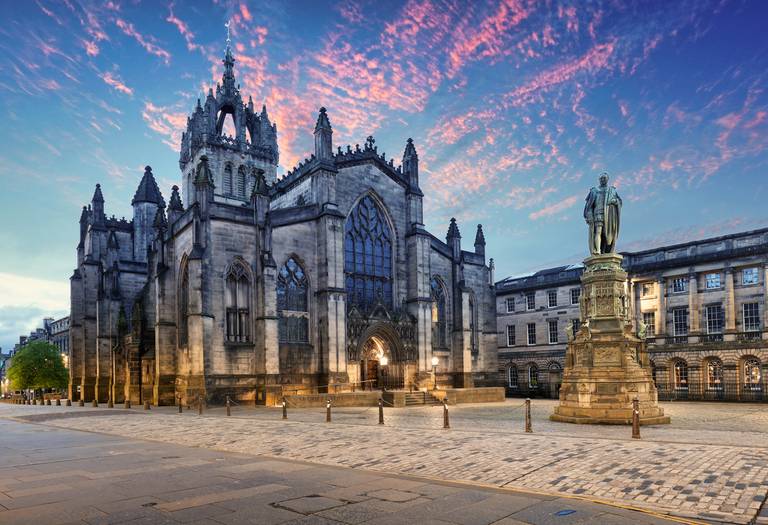 Historische gotische Kathedrale mit kunstvoller Architektur, großen Buntglasfenstern und einer Statue im Vordergrund, unter einem lebhaften rosa-blauen Himmel