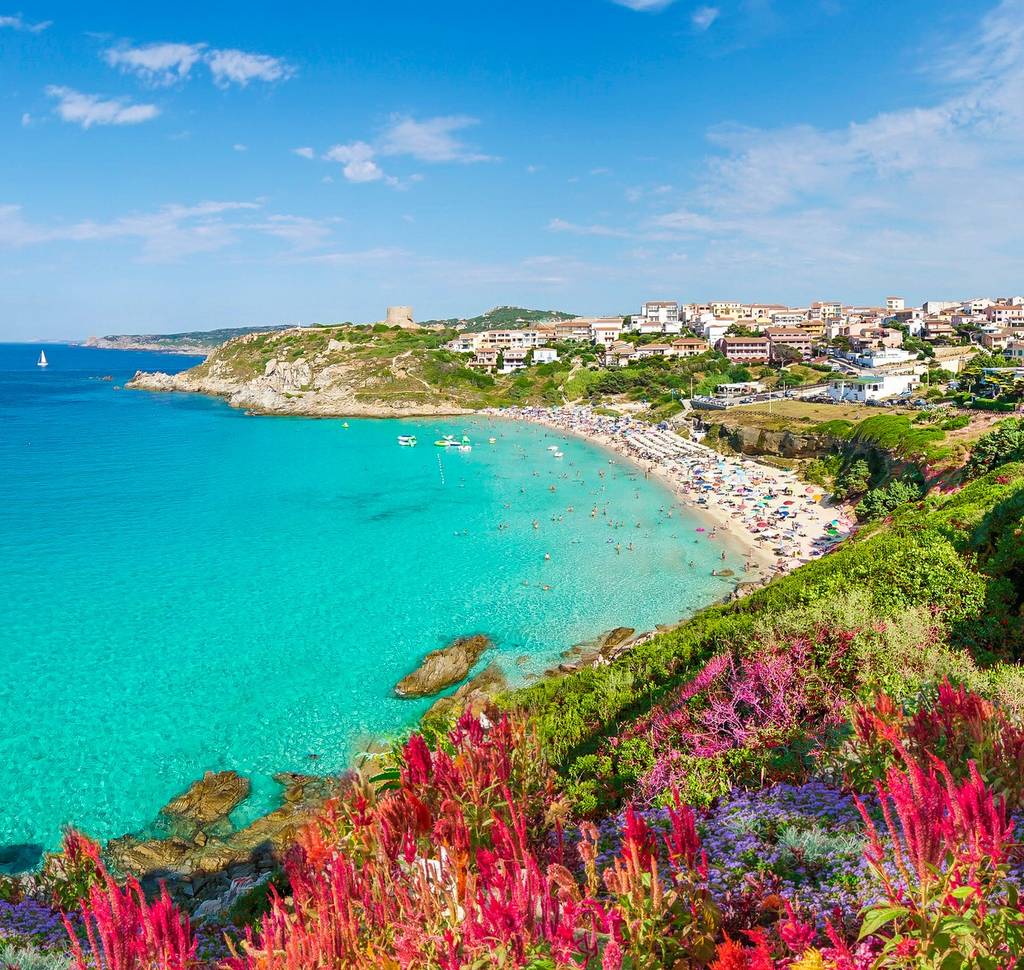 Coastal town with turquoise waters, sandy beach crowded with people, vibrant pink flowers in the foreground, and a clear blue sky.