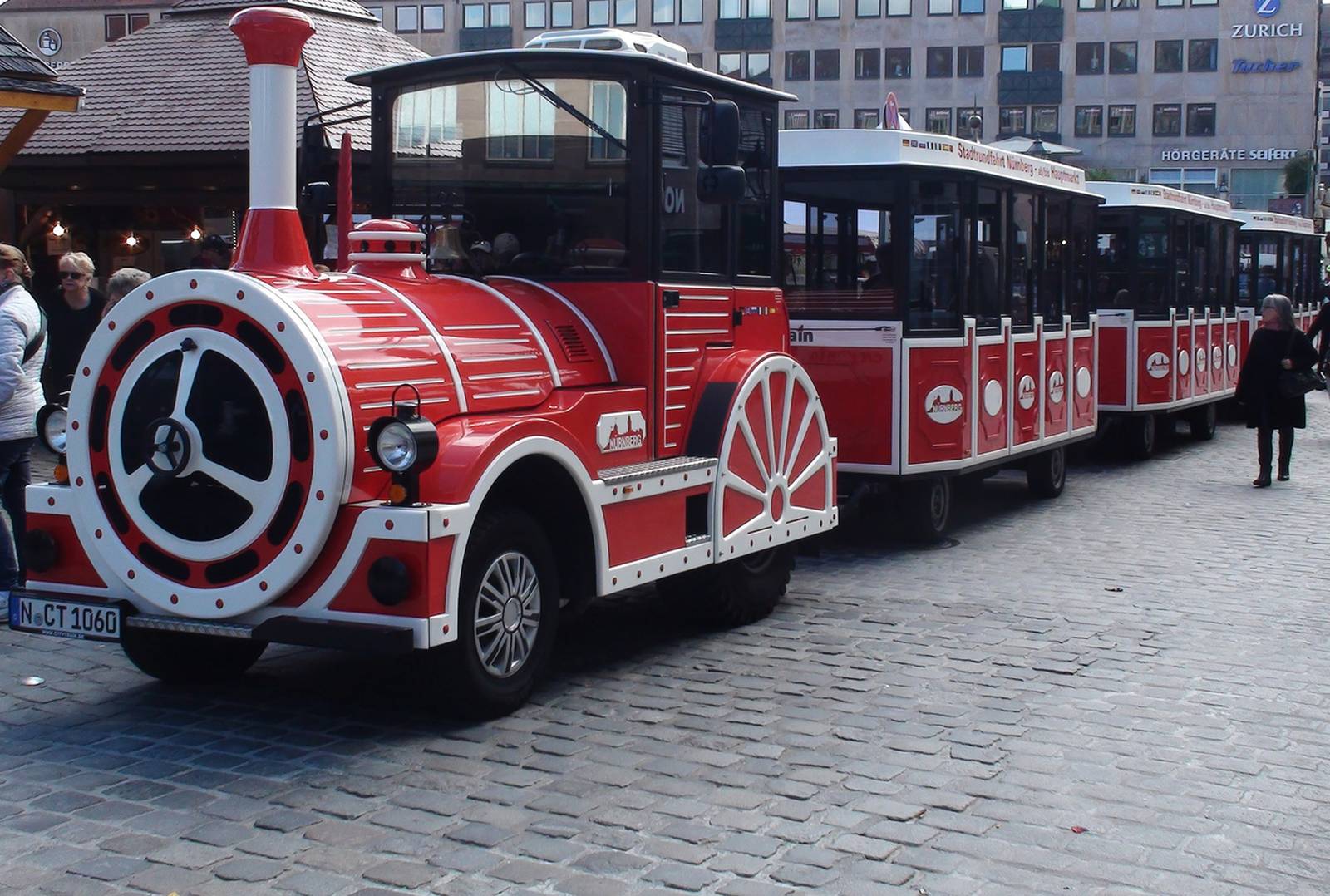 Ein rot-weißer Touristenzug mit mehreren Waggons steht auf einer Kopfsteinpflasterstraße in einem städtischen Gebiet