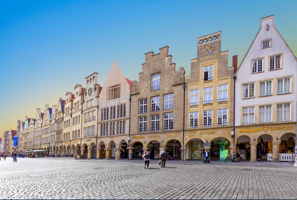 Historische europäische Straße mit farbenfrohen Giebelhäusern, Arkadengängen und einem Kopfsteinpflasterplatz unter klarem, blauem Himmel. In der Nähe flanieren Menschen