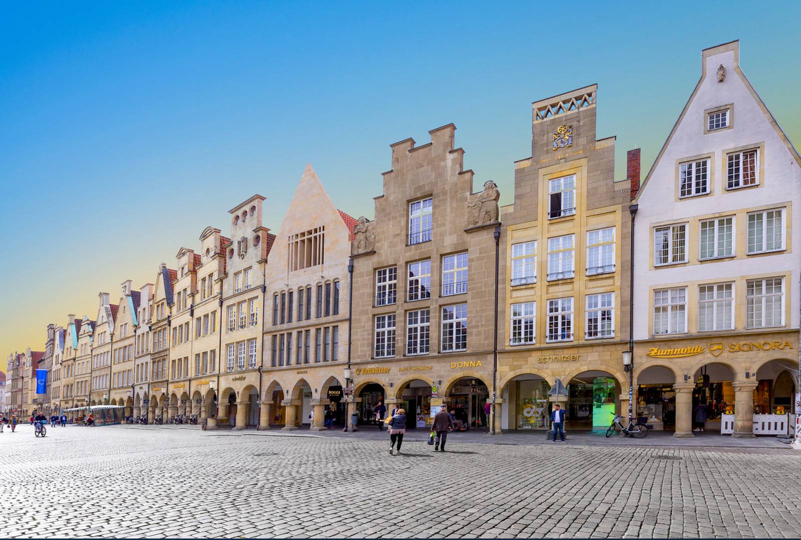 Historische europäische Straße mit farbenfrohen Giebelhäusern, Arkadengängen und einem Kopfsteinpflasterplatz unter klarem, blauem Himmel. In der Nähe flanieren Menschen