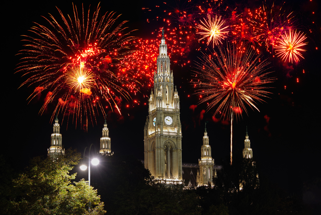 Colorful fireworks illuminate the night sky above a large, ornate, illuminated Gothic-style building surrounded by trees.