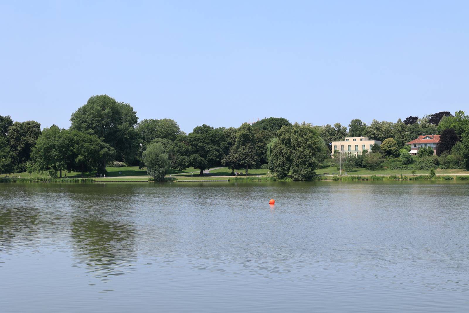Ruhiger See mit einem fernen, von Bäumen gesäumten Ufer, einer roten Boje im Wasser und einigen sichtbaren Gebäuden unter klarem, blauem Himmel