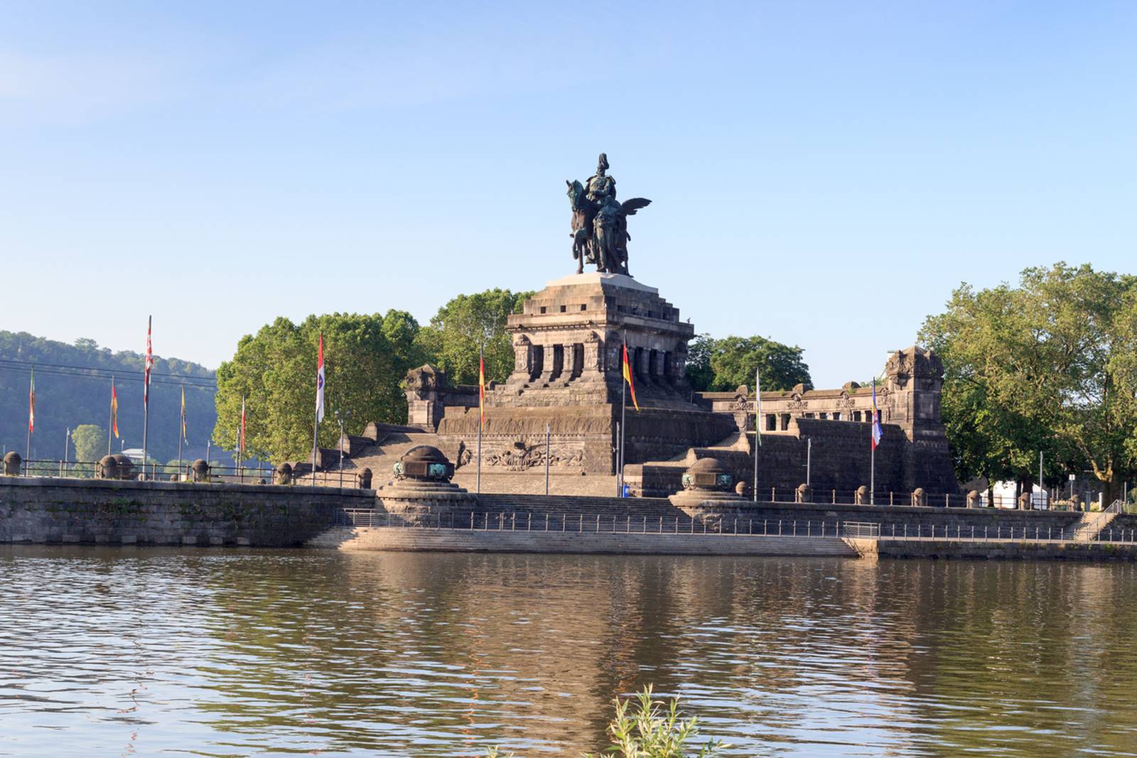 Reiterstatue auf einem großen steinernen Denkmal am Zusammenfluss zweier Flüsse, umgeben von Bäumen und Fahnen unter klarem, blauem Himmel