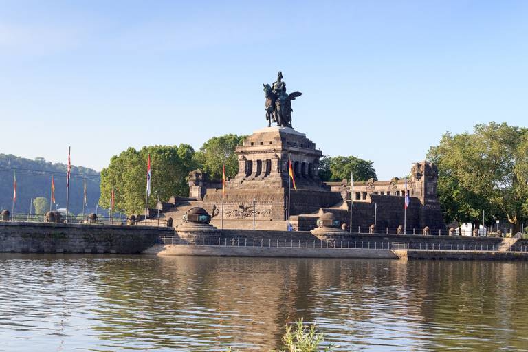 Reiterstatue auf einem großen steinernen Denkmal am Zusammenfluss zweier Flüsse, umgeben von Bäumen und Fahnen unter klarem, blauem Himmel