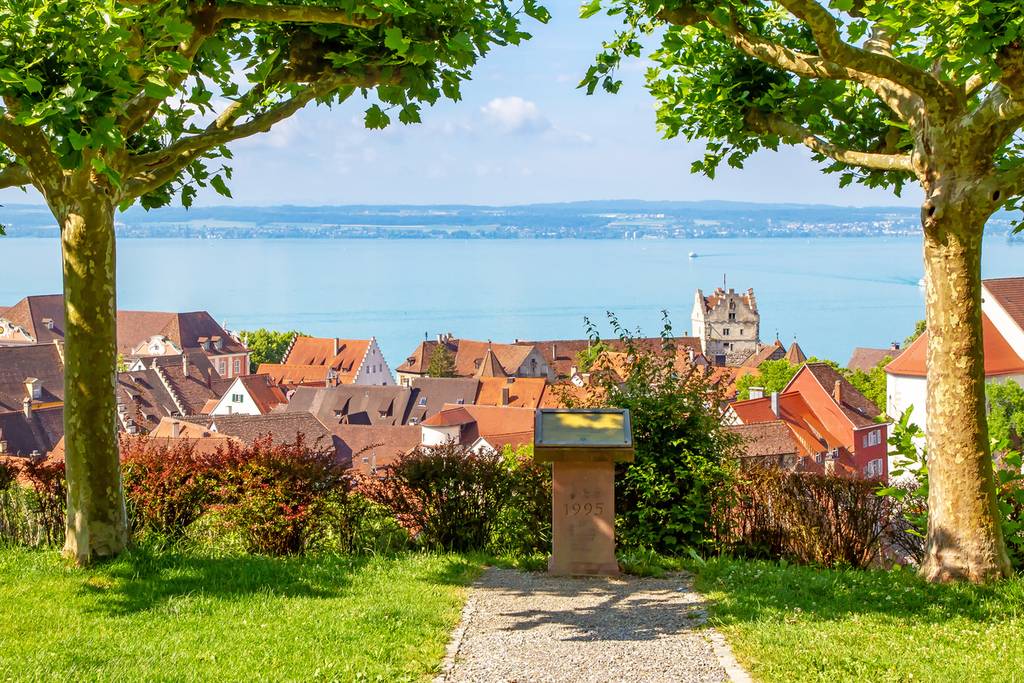 Path between two trees to an information plaque overlooking a red-roofed town and the Lake Constance under a clear sky.