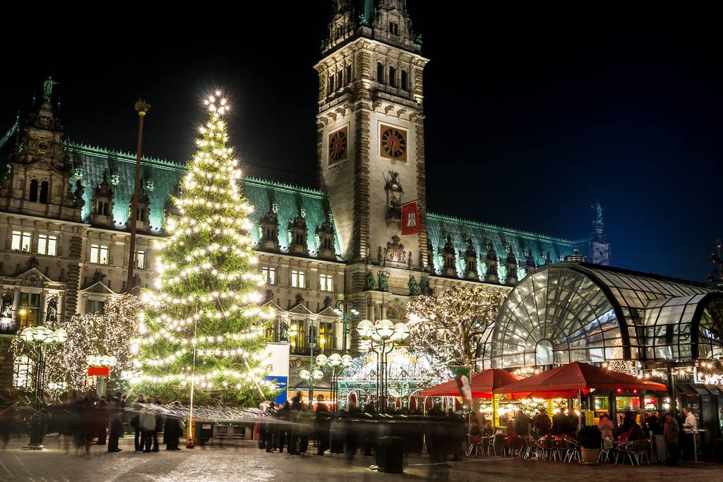 Festlicher Weihnachtsmarkt bei Nacht mit einem großen, beleuchteten Weihnachtsbaum, belebten Marktständen und einem prachtvollen historischen Gebäude im Hintergrund.