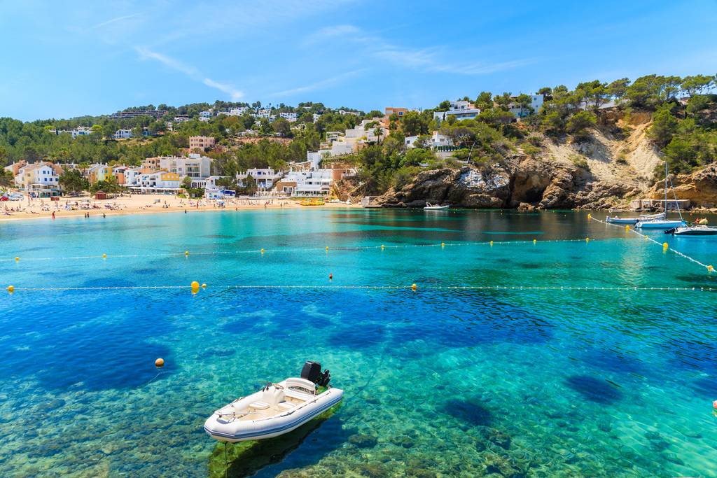 Vista panorámica de una bahía tranquila de aguas turquesa, con una pequeña barca flotando, playa de arena y casas en la ladera al fondo.