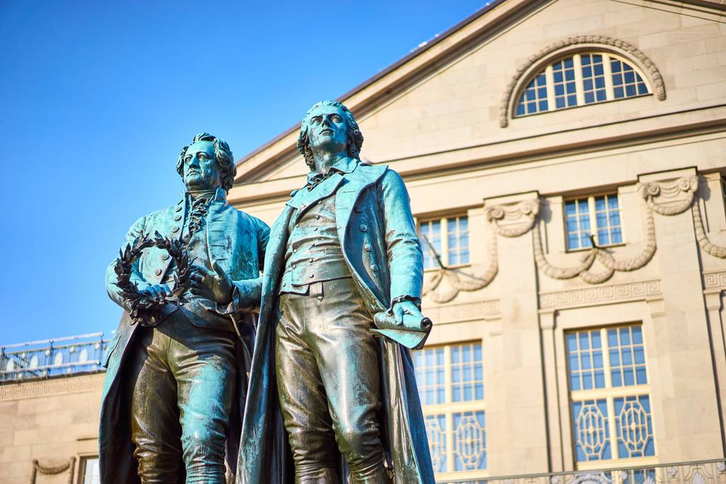 Bronzestatuen von zwei Männern in Kleidung aus dem 18. Jahrhundert stehen vor einem historischen Gebäude mit großen Fenstern und aufwendiger Steinornamentik.