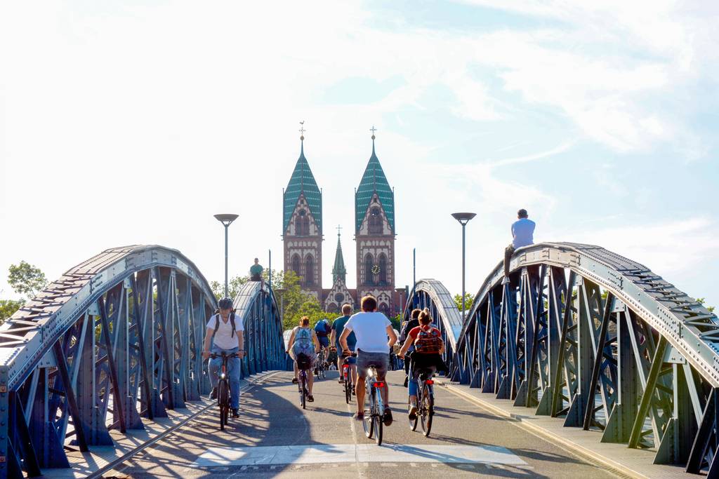 Radfahrer überqueren eine blaue Metallbrücke in Richtung einer entfernten Kirche mit zwei Türmen unter klarem Himmel