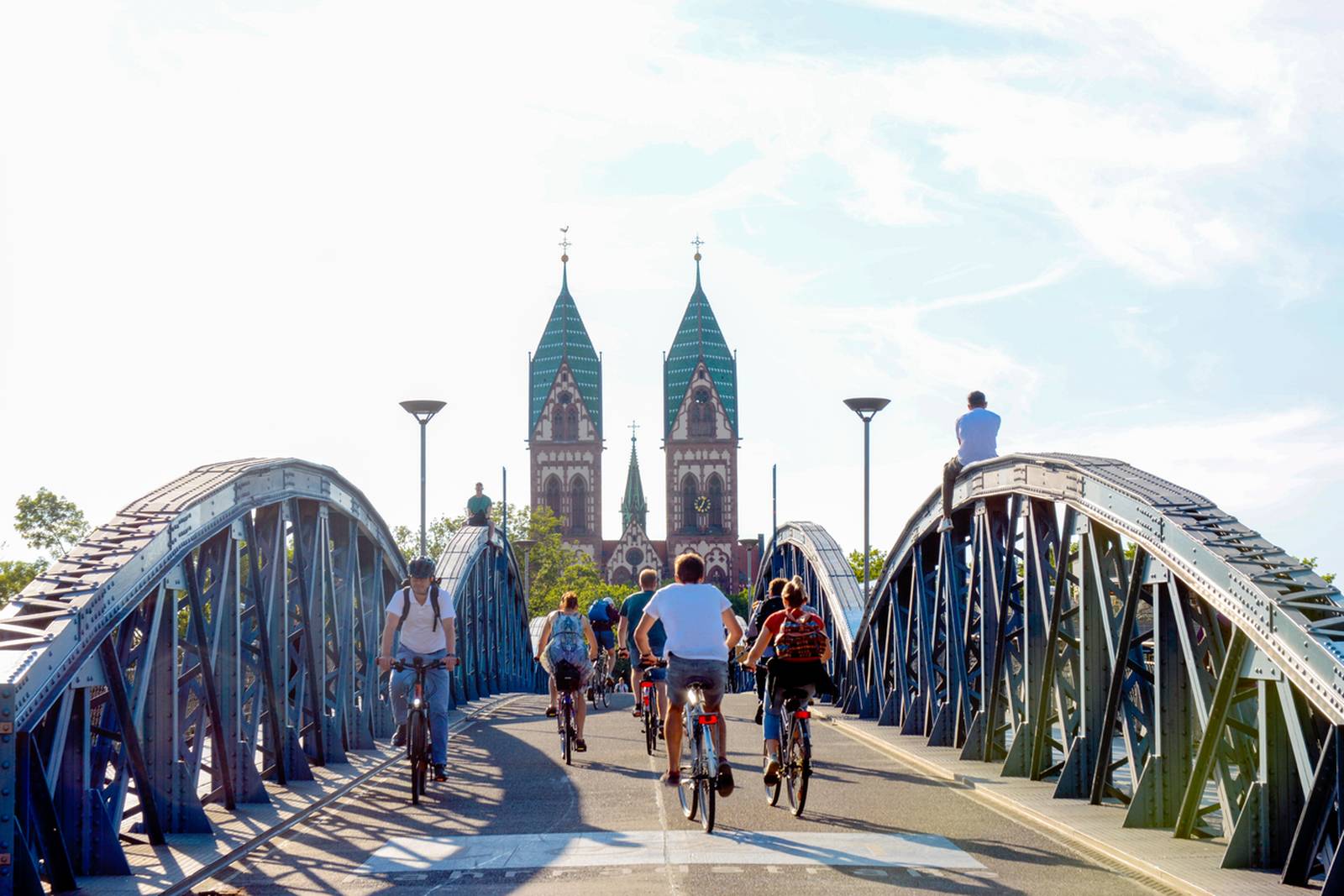Radfahrer überqueren eine blaue Metallbrücke in Richtung einer entfernten Kirche mit zwei Türmen unter klarem Himmel