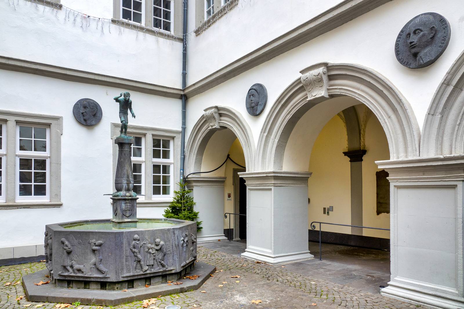 Historischer Innenhof mit einem bogenförmigen Eingang, dekorativen Plaketten und einem zentralen Brunnen mit einer kleinen Statue