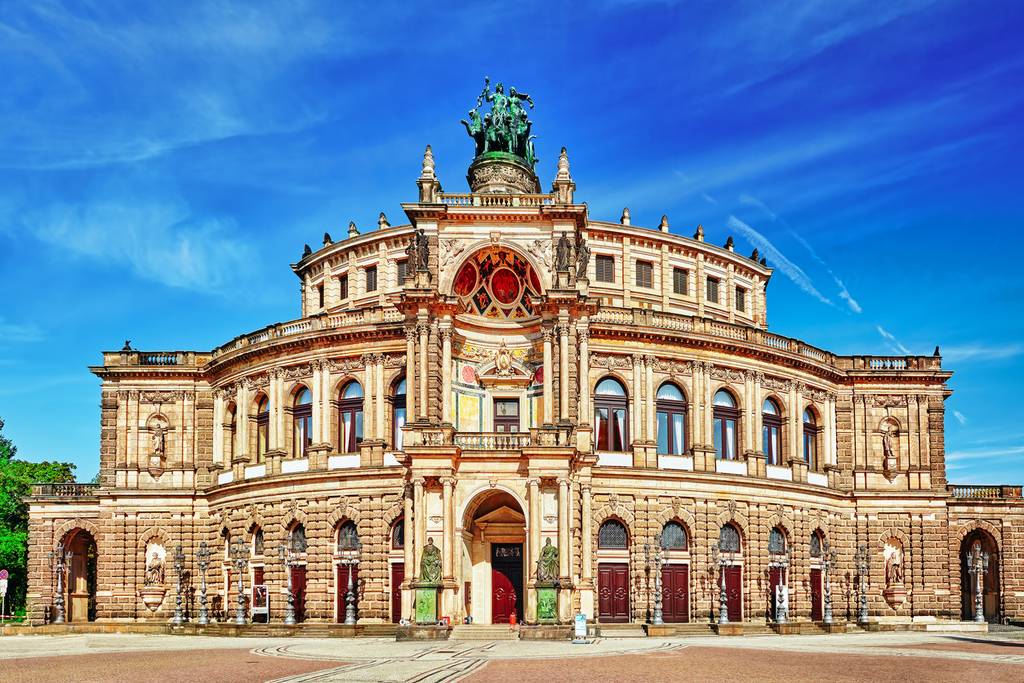 Ornate historic opera house facade with arched windows, decorative statues and a sculpted group atop under a bright blue sky.