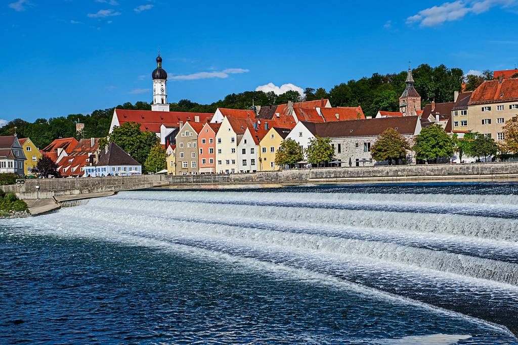 Europäische Stadt am Fluss mit bunten Giebelhäusern und Kirchturm hinter einem gestuften Wehr, das über den Fluss stürzt, unter blauem Himmel.