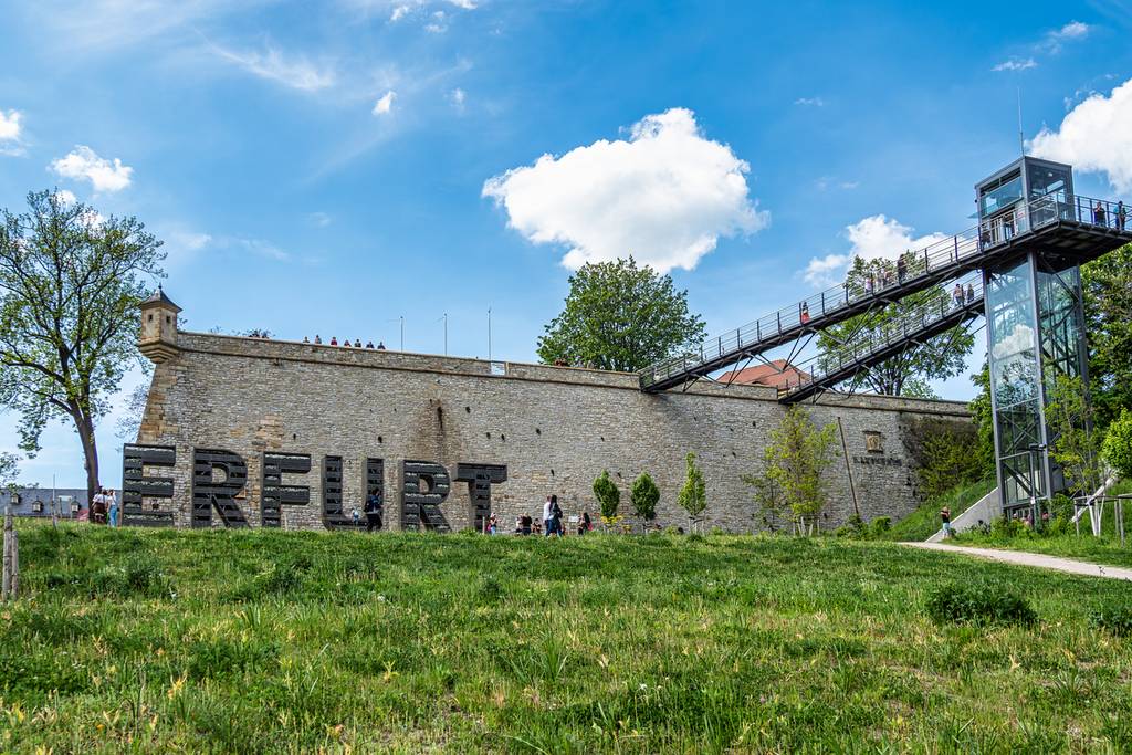Historische Steinmauer in Erfurt mit großen Buchstaben „ERFURT“ im Vordergrund, umgeben von Grünflächen und einem gläsernen Aufzug auf der rechten Seite
