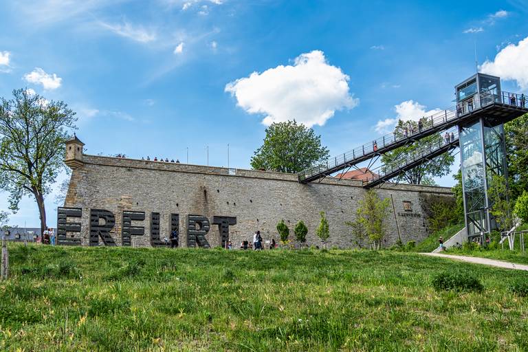 Historische Steinmauer in Erfurt mit großen Buchstaben „ERFURT“ im Vordergrund, umgeben von Grünflächen und einem gläsernen Aufzug auf der rechten Seite