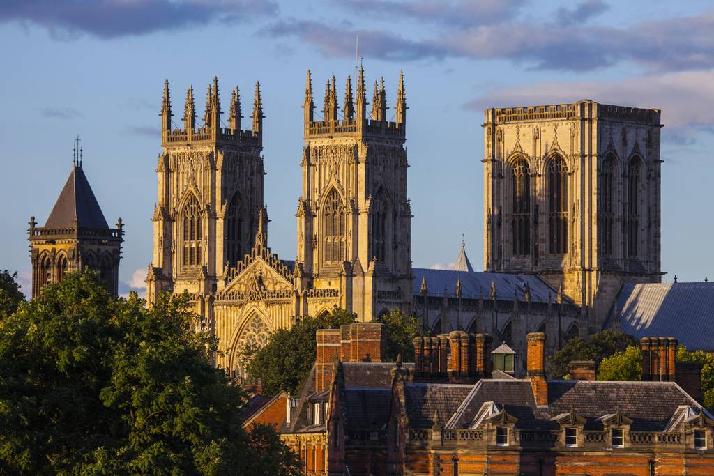 Gothic cathedral of York with tall, intricate towers and spires, surrounded by trees and rooftops under a partly cloudy sky.