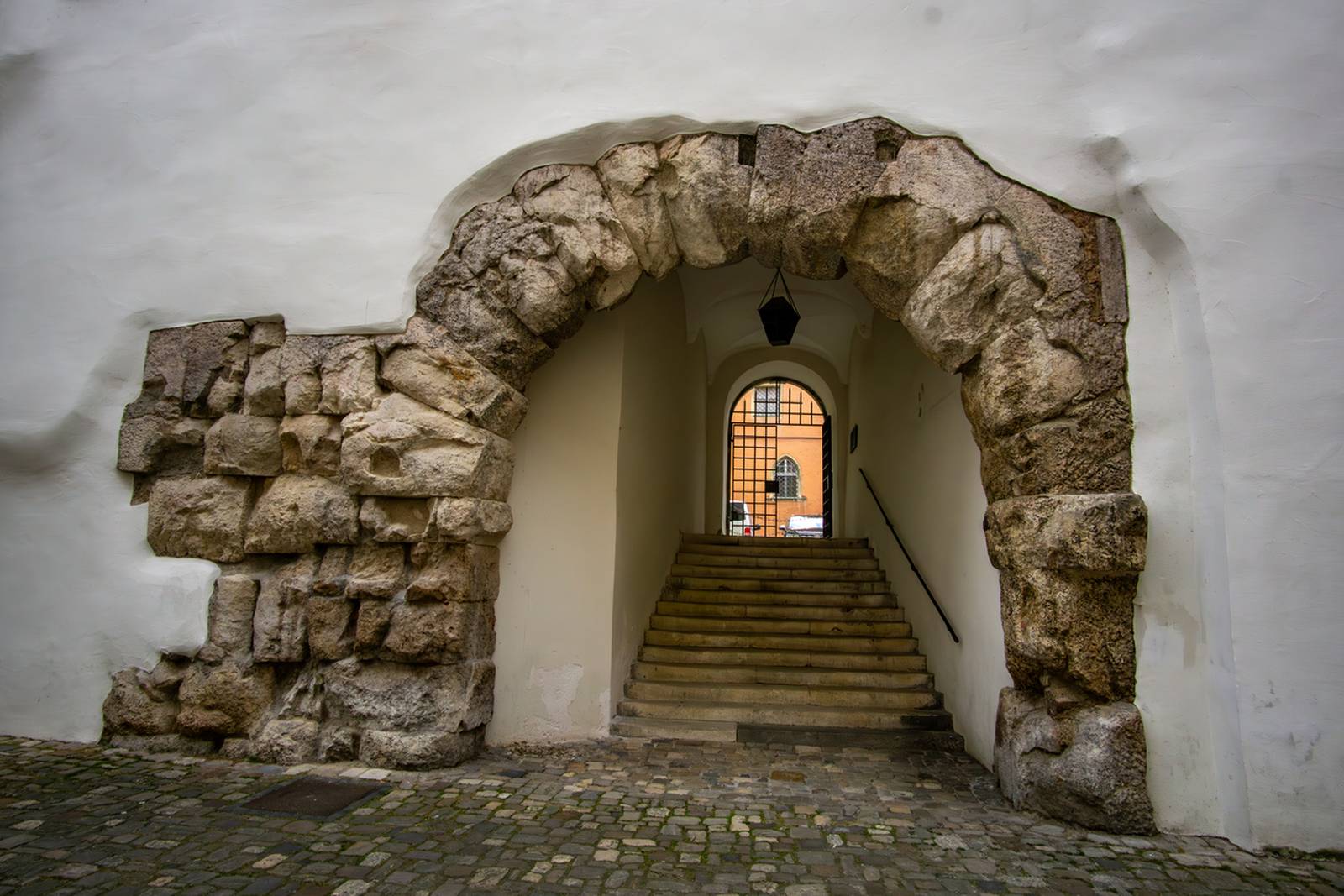 Steinernes Torbogen führt zu einer Treppe mit einer Laterne darüber, eingebettet in eine weiße Wand, mit Kopfsteinpflaster im Vordergrund