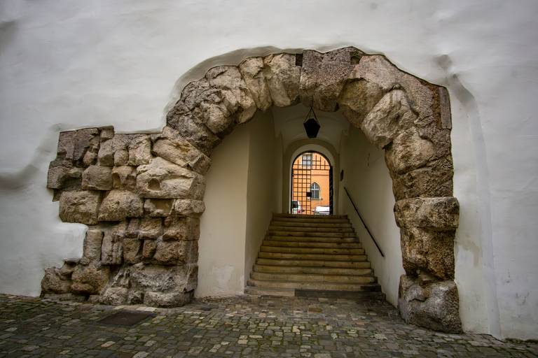Steinernes Torbogen führt zu einer Treppe mit einer Laterne darüber, eingebettet in eine weiße Wand, mit Kopfsteinpflaster im Vordergrund