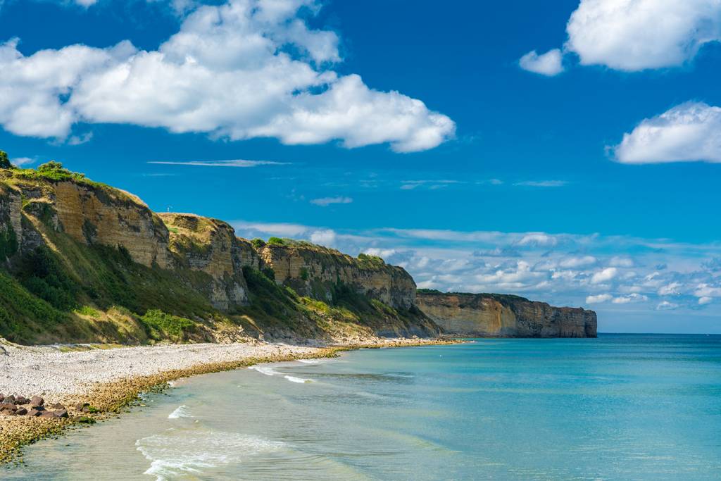 Vue panoramique sur un littoral rocheux aux falaises abruptes, aux eaux turquoise cristallines et au ciel bleu parsemé de quelques nuages.